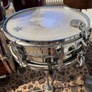 Kovový snare drum stříbrné barvy s bílým membránou, která vykazuje známky opotřebení a znečištění. Na membráně je viditelné logo s nápisem REMO. Nást…