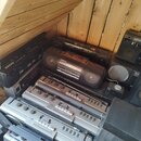 Sestava několika černých a stříbrných přenosných radiokasetových přehrávačů naskládaných na sebe v dřevěném prostoru. Na zařizení jsou viditelné nápi…