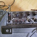Kytarový kombozesilovač značky JOYO, model DC-15, v černé barvě s texturovaným povrchem. Ovládací panel obsahuje nápisy POWER ON, AMP (s voličem BLUE…