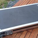 Černý transportní kufr s hliníkovým rámem a kovovými rohy, položený na dřevěném stole. V pravém horním rohu je čtvercový štítek s logem „GR“. Kufr má…