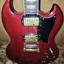 Červená elektrická kytara s černým pickguardem a dvěma humbuckery se zlatými kryty. Na hmatníku jsou vidět bílé trojúhelníkové vložky a na těle se na…