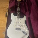 Bílá elektrická kytara typu Stratocaster v otevřeném tvrdém kufru s vínovým polstrováním. Na hlavě hmatníku je nápis Fender. Kytara má tři snímače, b…