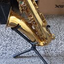 Zlatý saxofon umístěný na černém kovovém stojanu. Nástroj vykazuje známky používání v podobě drobných škrábanců a znečištění povrchu. Na těle nástroj…