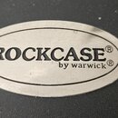 Oválný kovový štítek s nápisem ROCKCASE® a pod ním menším písmem by warwick®. Štítek je připevněn na černý, texturovaný povrch. Povrch kovového prvku…