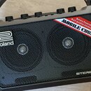 Černý přenosný stereo zesilovač s kovovou mřížkou přes reproduktory. Na levé straně je bílé logo Roland a vpravo dole nápis STEREO. V horní části je…