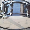 Černý snare drum s kovovým obručováním a ladicími šrouby. Na spodní straně je vidět bílá membrána s logem v podobě zvířete. Nástroj je umístěn na dře…