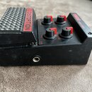 Černý efekt pedál s integrovaným spínacím pedálem. Na horní části jsou čtyři černé oválné knoflíky s červenými značkami a nápisy DISTORTION, TUNE, BA…