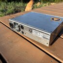 Stříbrné kovové zařízení v hranatém šasi s ventilátorem na zadní straně. Na horní části je kulaté žluté logo s nápisem "SURE" a "POWER SUPPLY". Na za…