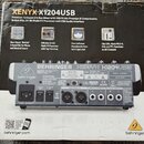Fotografie znázorňuje kartonové balení míchacího pultu. Na krabici je nápis XENYX X1204USB a logo BEHRINGER. V horní části je text Premium 12-Input 2…