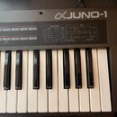 Černý klávesový syntetizátor s bílými a černými klávesami. Na horním panelu je nápis JUNO-1 doprovázený logem. Panel obsahuje tabulky s popisky WAVEF…