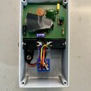 Fotografie ukazuje vnitřek bílého kovového krabičky s elektronickým obvodem na zelené desce. Na desce jsou připájeny různé součástky, včetně kondenzá…