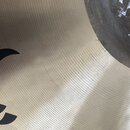 Detailní záběr na povrch kovového činele zlatavé barvy s viditelným koncentrickým rýhováním. V levé části je vidět fragment černého loga nebo nápisu.…