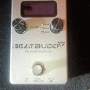 Stříbrný efekt pedál s nápisy BEATBUDDY MINI a Personal Drummer. Zařízení má digitální displej, dva ovládací knoflíky s popisy VOLUME a GENRE SONG TE…