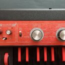 Červený syntetizátor s černým šasi a klávesami. Na horním panelu je bílý nápis KORG microKORG XL synthesiser vocoder. Jsou zde vidět ovládací knoflík…