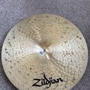 Kruhový činel z mosazi s viditelným kovaným povrchem a drobnými tmavšími skvrnami. V dolní části je černý nápis Zildjian a nad ním nečitelný podpis.…