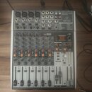 Šedý mixovací pult značky behringer s nápisem 24-BIT MULTI-FX PROCESSOR. Zařízení obsahuje několik vstupních kanálů s posuvníky, červené a modré ovlá…