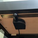 Detailní pohled na černý plastový mechanismus s úchytem, který je připevněn k dřevěné desce. Nad mechanismem je vidět kovová lišta a černý lakovaný p…