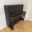 Tmavě hnědé pianino s dřevěnou texturou umístěné v rohu místnosti na světlé podlaze. Povrch nástroje vykazuje známky opotřebení, zejména v podobě odř…