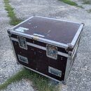Černý transportní case s hliníkovými hranami a rohy. Povrch je z texturovaného materiálu s viditelným opotřebením a několika bílými nálepkami. Na pře…