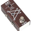 Hnědý kovový pedál s bílým nápisem ABY Channel Switch. Na horní straně je přepínač s popisky A/B-V a A&B-V, pod ním text A-Y:RED B-V:BLUE A&B-V:PINK…