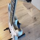 Elektrická kytara bílé barvy umístěná na černém stojanu. Na hmatníku jsou viditelné vložky s ptačím motivem. Povrch kytary vykazuje výrazné opotřeben…