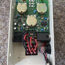 Fotografie zachycuje vnitřek elektronického zařízení v bílém kovovém šasi. Na zelené základní desce jsou umístěny tři zlatavé kondenzátory s nápisy „…