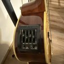 Detailní pohled na ovládací panel akustické kytary s hnědým povrchem. Černý plastový panel obsahuje čtyři posuvníky s popisky BASS, MIDDLE, TREBLE a…