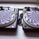 Dvě černé turntable gramofony s instalovanými hracími rameny. Tallyře jsou zakryty dekorativními podložkami s fialovo-bílou spirálovitou grafikou, na…
