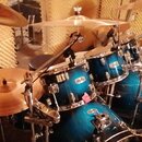 Modrá bicí souprava s kovovým hardwarem. Na jednom z činelů je viditelný nápis Paiste. Souprava obsahuje basový buben, několik tomů a činely umístěné…