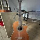 Akustická kytara s nylonovými strunami v přirozené dřevěné barvě, umístěná na černém stojanu. Kytara má tvar cutaway a ozdobný lem kolem otvoru rezon…