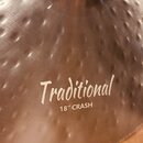 Detailní záběr na kovový činel stříbrné barvy s kovaným povrchem (hammered). Na povrchu je bílý nápis „Traditional“ a pod ním text „18" CRASH“. Povrc…