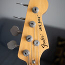 Fotografie zachycuje hlavu basové kytary z přírodního světlého dřeva. Na povrchu jsou černé nápisy „Fender“, „JAZZ BASS“ a menší texty „MADE IN JAPAN…