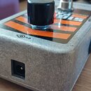Kovový efekt pedál stříbrné barvy s hrubým povrchem. Na horní straně je černý oválný knoflík a oranžovo-černý pruhovaný štítek s neúplně viditelným n…