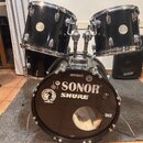 Černá bicí souprava tvořená dvěma tommy a basovým bubnem. Na basovém bubnu jsou viditelné nápisy SONOR, SHURE, METALO a bern, dále logo National Drum…