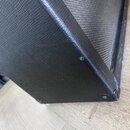 Detailní pohled na roh černého reproduktorového boxu potaženého texturovaným vinylen. Na hranách jsou vidět kovové ochranné rohy a v místě spoje je p…