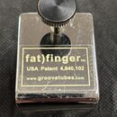 Kovový předmět stříbrné barvy s hranatým tvarem a vrutem s knurlovaným knoflíkem. Na povrchu je nalepený štítek s nápisy (fat)finger™, USA Patent 4,8…