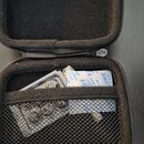 Otevřený černý zipový pouzdro s vnitřním polstrováním a síťovou kapsou. V kapse jsou umístěny drobné předměty, včetně stříbrného kovového příslušenst…
