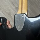 Detail zadní strany černé kytary v místě spoje krku s tělem. Kovový kryt krku je připevněn čtyřmi šrouby a nese vyryté sériové číslo 712794, podpis a…