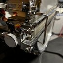 Detailní pohled na chromovaný mechanismus pedálu bicího bubnu. Je vidět kovový táh, pružina a nastavovací šrouby s rýhovaným knoflíkem. Povrch je les…