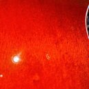 Detailní záběr na červený lakovaný povrch hudebního nástroje s viditelnou strukturou dřeva. V horní části je vidět okraj kulatého otvoru. Uprostřed p…
