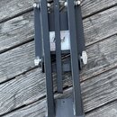 Černý kovový mechanismus pedálu, pravděpodobně součást bicího pedálu. Uprostřed je viditelný štítek s nápisem "Pearl". Povrch vykazuje mírné známky o…