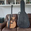 Akustická kytara s cutawayem, přírodní barvou korpusu a tmavým hmatníkem. Na hlavě nástroje je viditelné logo "Tanglewood". Vedle kytary stojí černý…