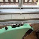 Detailní pohled na zadní část světle zelené elektrické kytary. Jsou vidět čtyři černé potenciometry a jeden přepínač s červeným knoflíkem. Na zadní s…