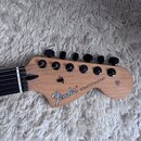 Fotografie zachycuje hlavu elektrické kytary z přírodního dřeva s šesti černými ladičkami. Na dřevě je uveden nápis Fender® STRATOCASTER® a v pravé č…