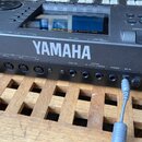 Černý elektronický klávesový nástroj s bílým nápisem YAMAHA na přední straně. Na zadním panelu jsou viditelné konektory s popisky OUT-B, OUT-A, MIDI,…