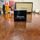Na dřevěném stole stojí černá čtvercová krabice s bílým nápisem Ibanez Bass Tube Screamer TS9B. V pozadí je vidět béžový kytarový kombozesilovač s ná…