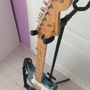 Elektrická kytara s modrými doplady a světle hnědým hnedem z dřeva, umístěná na černém stojanu. Na zadní straně hlavy je nápis „Fender® STRATOCASTER®…