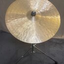 Zlatý kovový činel s kovaným povrchem, umístěný na chromovaném stativu. Na povrchu je vidět kurzivní nápis "Paiste". Činel vykazuje mírné známky použ…