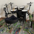 Elektronická bicí souprava s černým rámem a pět verifyingmi padů. Na modulu je nápis Roland, na pedálech značka Roland. Součástí je černý polstrovaný…