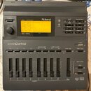 Šedý digitální modul Roland SC-155 Sound Canvas. Zařízení má podsvícený displej s textem „01 001 Piano 1“, řadu tlačítek a osm posuvníků s označením…